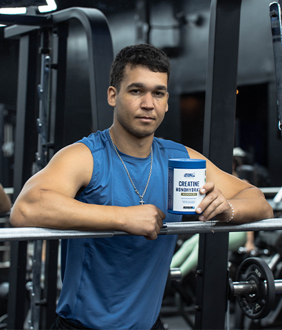 Hombre en gimnasio sosteniendo envase de suplemento junto a barra de pesas